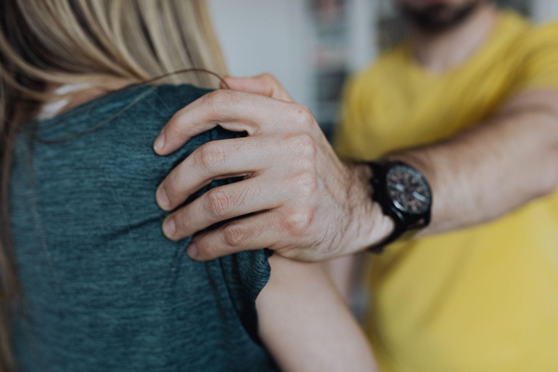 Hand resting on another person’s shoulder in a close indoor scene, with a yellow shirt in the background