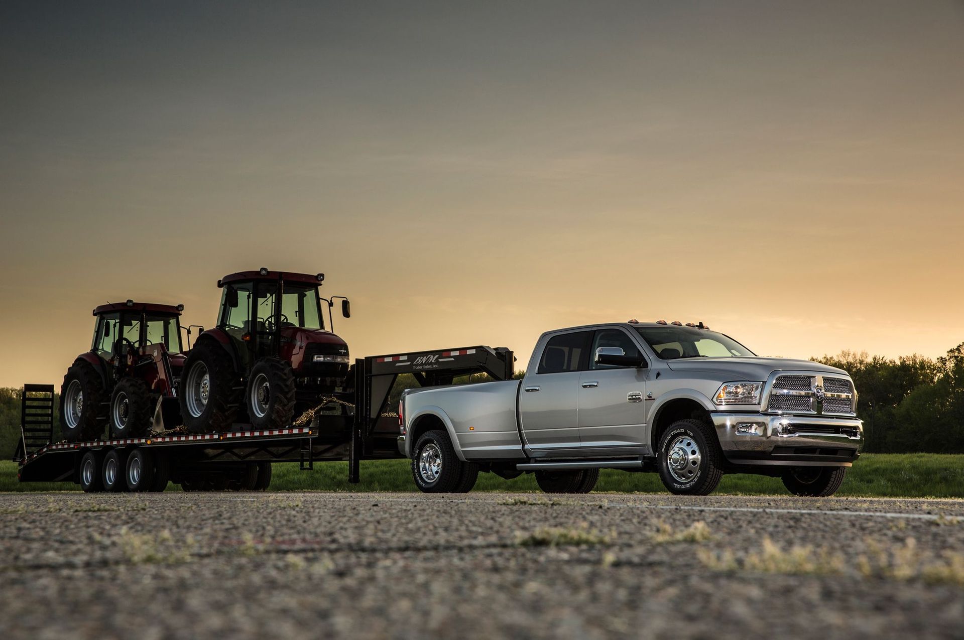 Silver pickup truck hauling two tractors on a flatbed trailer at sunset.