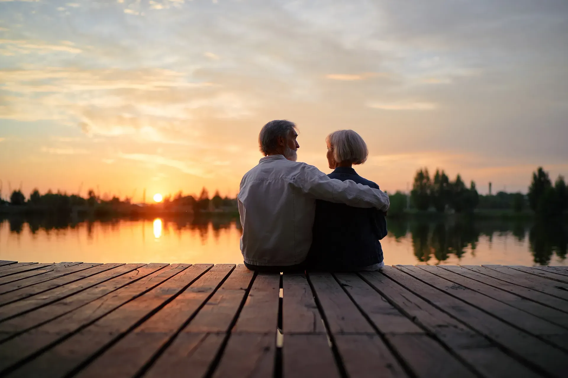 An elderly couple is sitting on a dock watching the sunset over a lake.