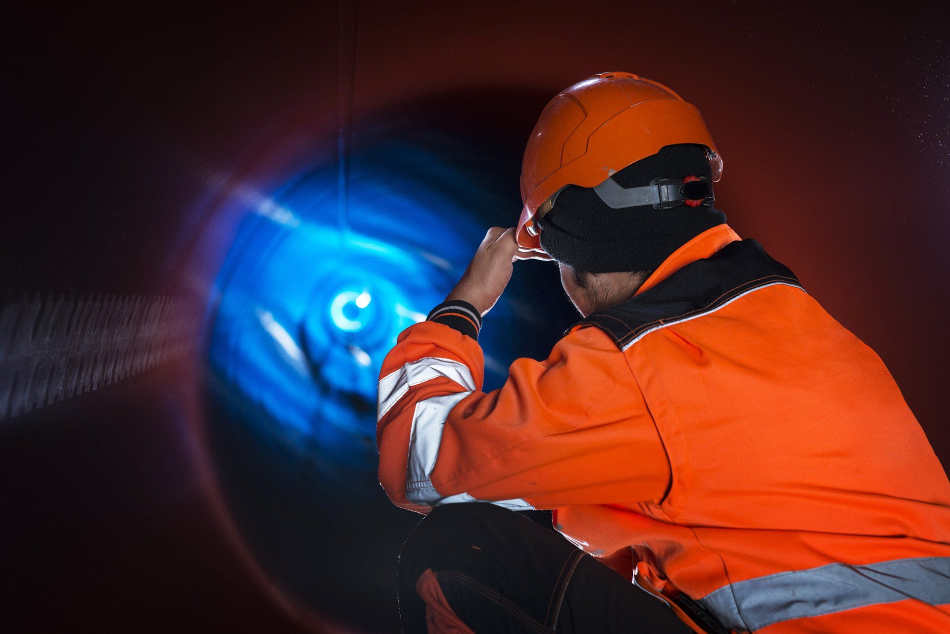 A worker in orange high-visibility gear and a hard hat kneeling inside a dark pipe, looking toward a glowing blue light.