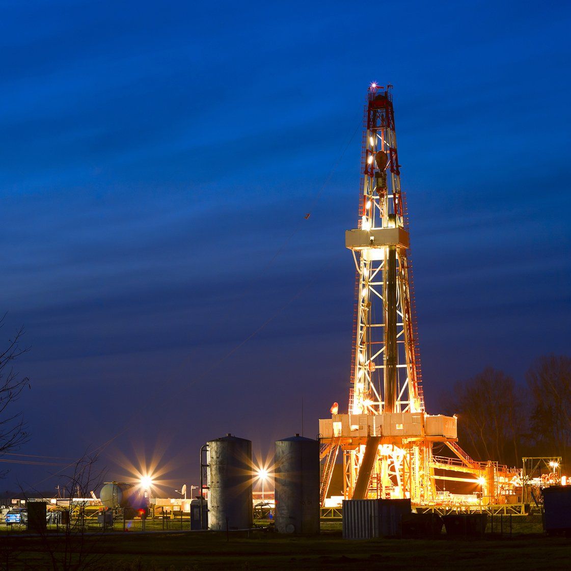 An oil drilling rig illuminated at night against a deep blue dusk sky, with storage tanks in the foreground.