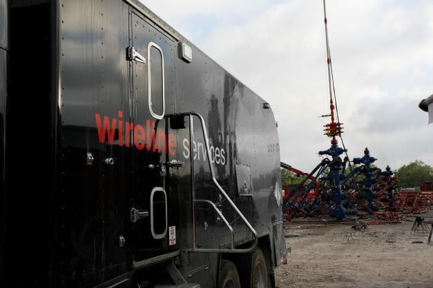 A black wireline services truck parked near an oil wellhead structure under a cloudy sky.