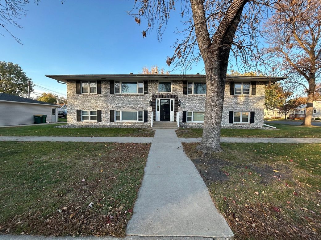 Two-story stone building with black shutters, sidewalk leading to the entrance, a tree to the right, and grass.