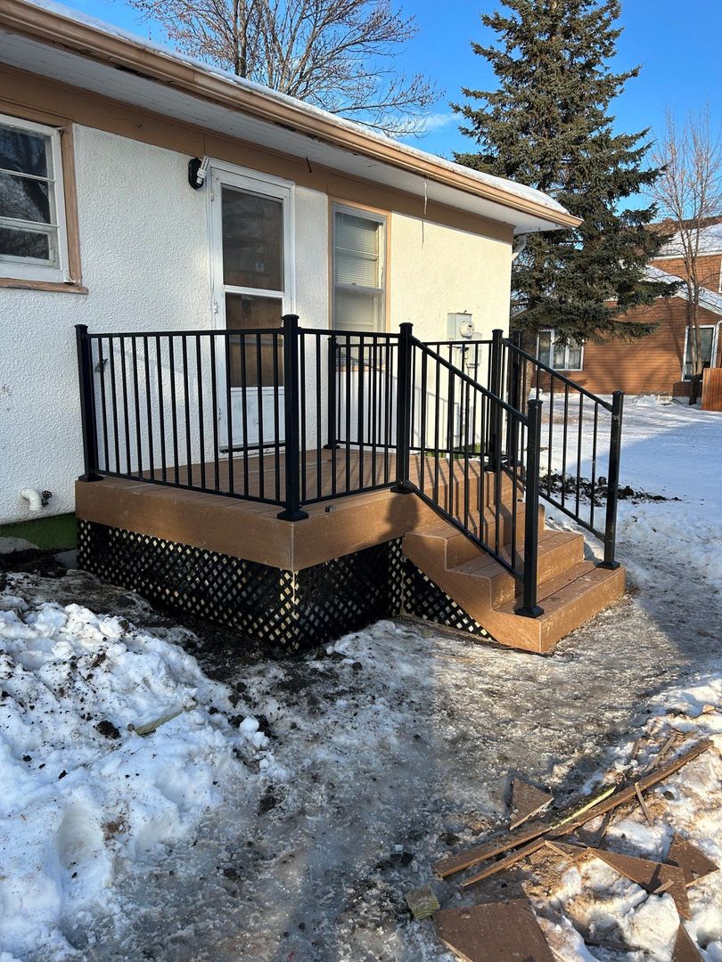 Brown deck with black railing and steps against a white house, snow on the ground.