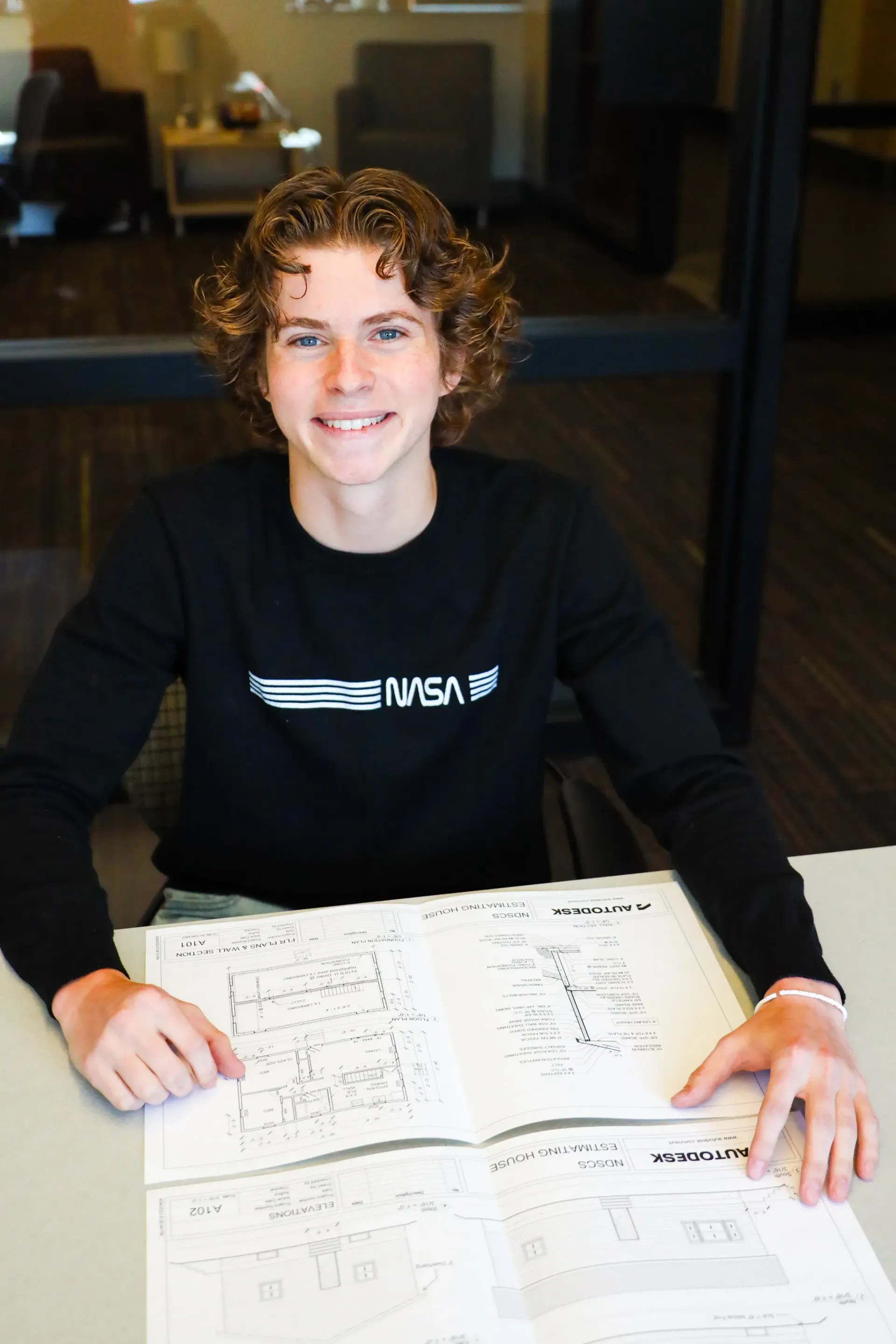 Young person with curly hair smiles, holding diagrams, wearing a black NASA shirt. Seated at a table.