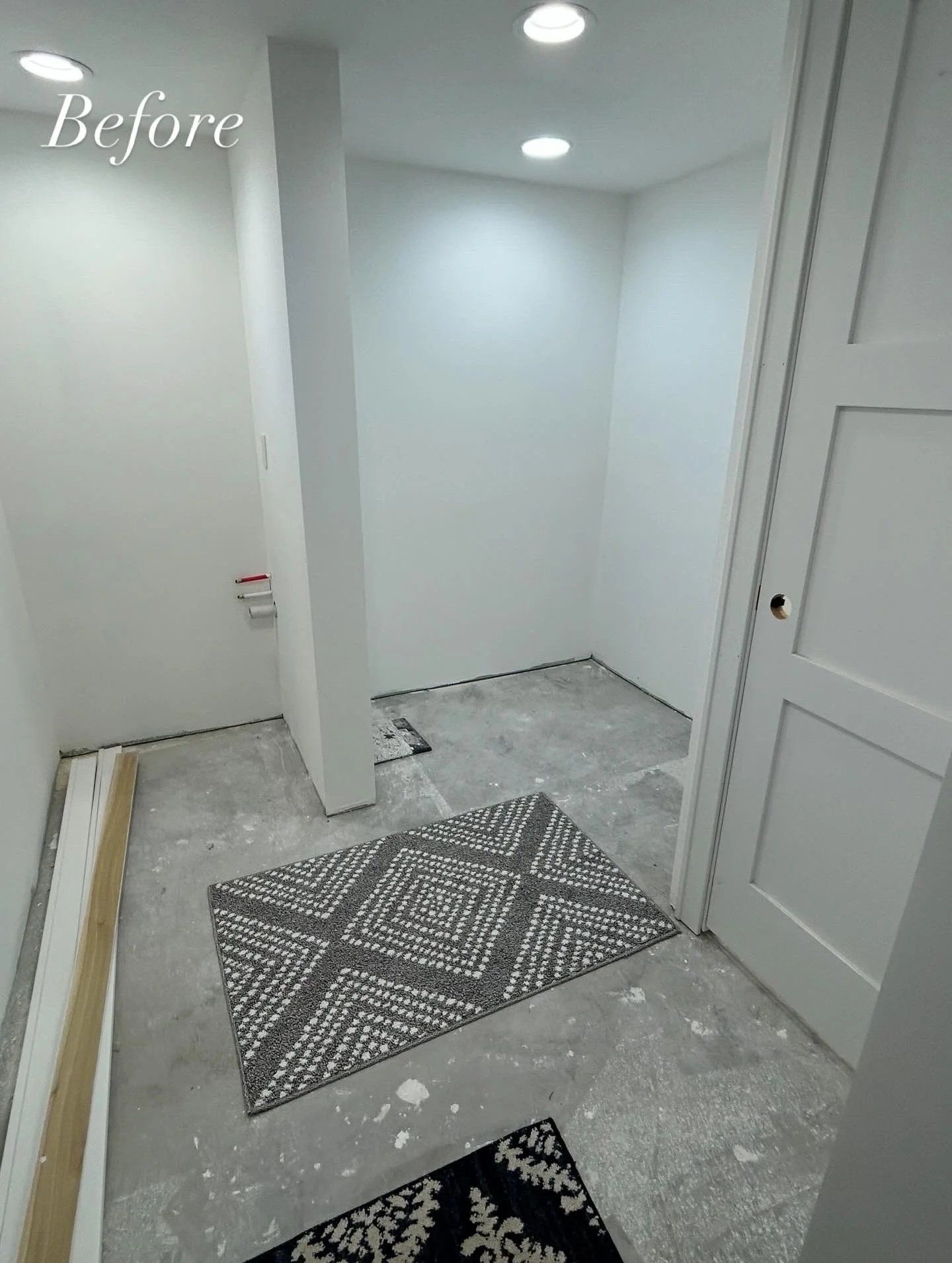 An empty white room with gray flooring, a door, and overhead lighting. A rug is on the floor.
