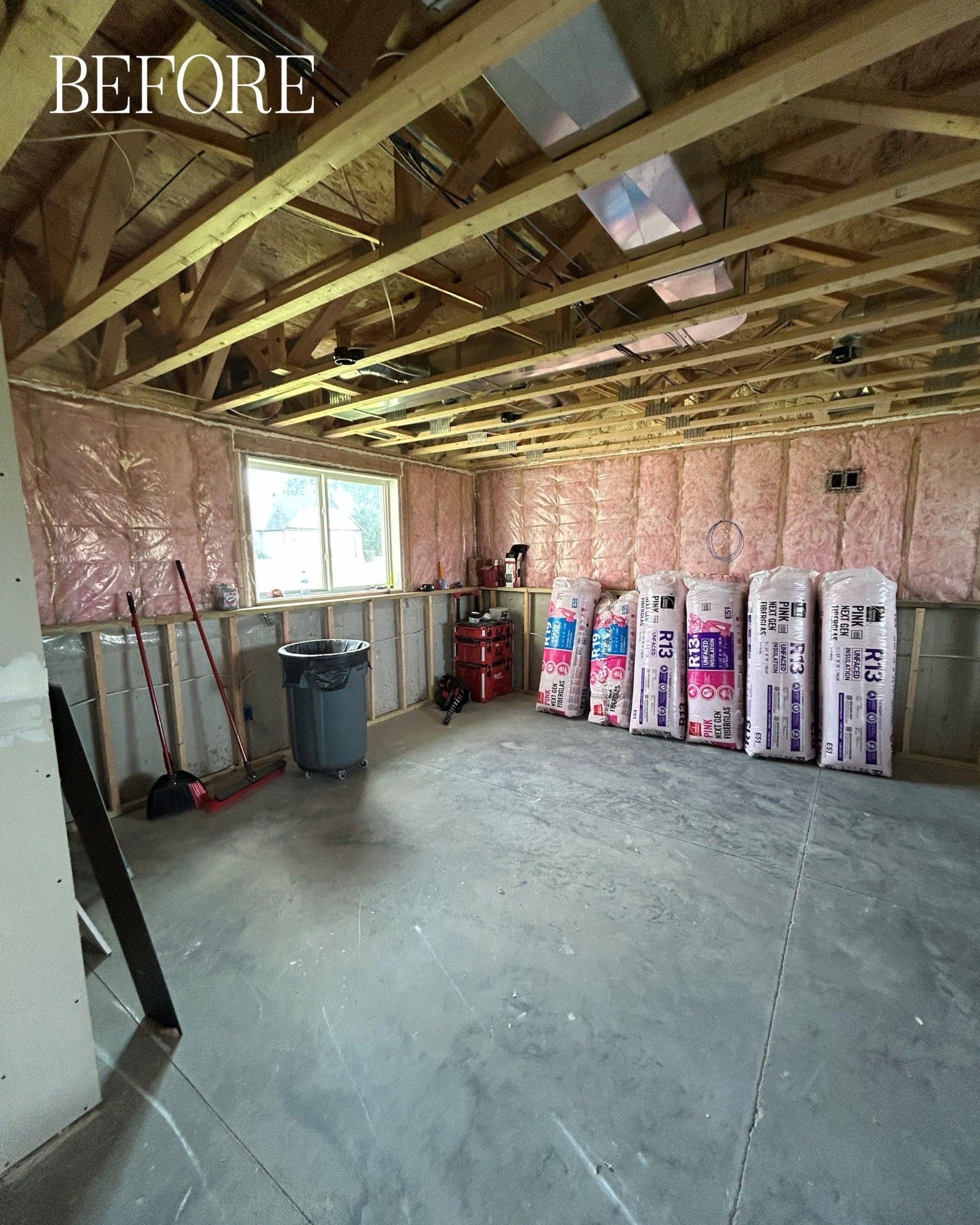 Unfinished basement before renovation. Pink insulation, exposed beams, concrete floor, bags of insulation. 