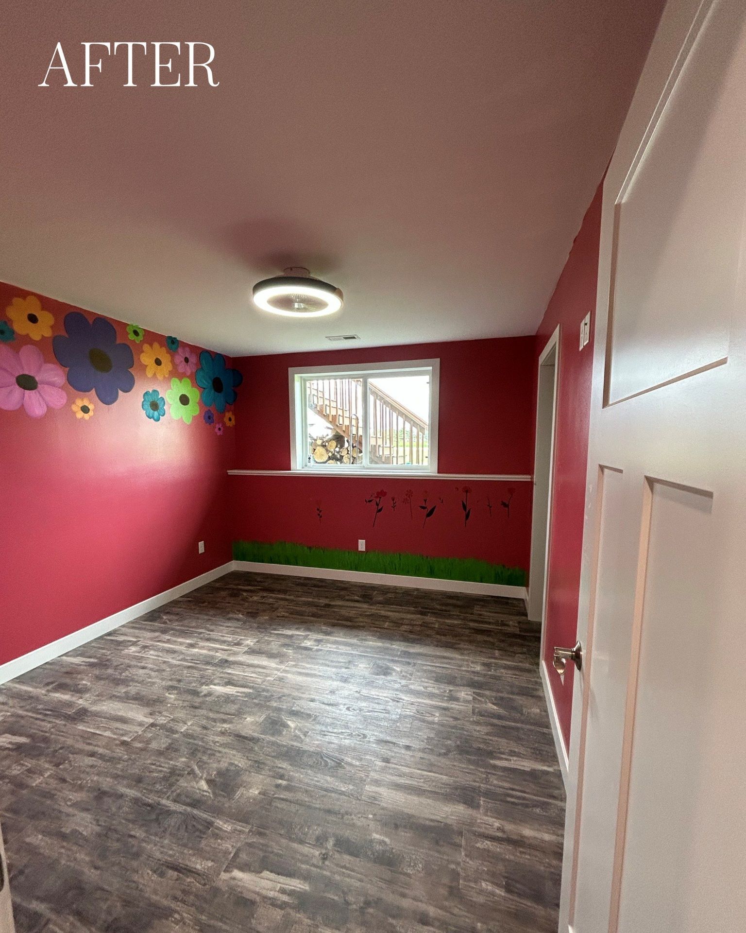Red-walled playroom with floral wall art, window, and brown flooring; lit by a ceiling light.