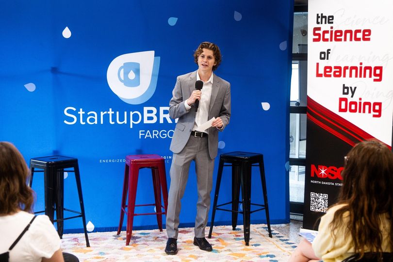 Man in a gray suit speaks at a StartupBRE Fargo event, holding papers. Blue backdrop, logo, and audience.