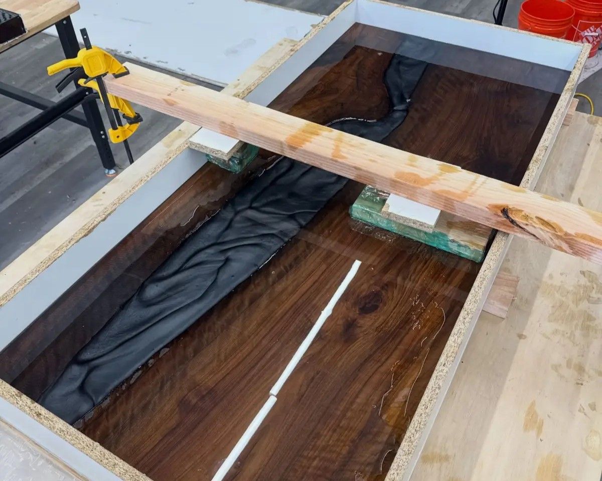 Wooden coffin lined with black fabric, supported in a workshop or construction area with clamps and boards.