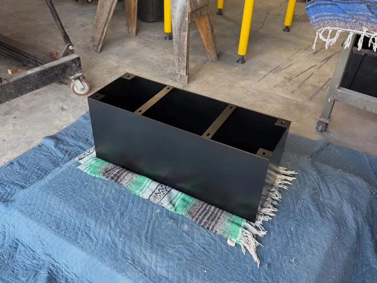 Black rectangular box on a blue tarp in a workshop, with open top compartments and yellow stools in the background