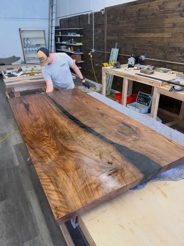Man sanding a large live-edge wooden slab in a workshop with tools and workbenches in the background