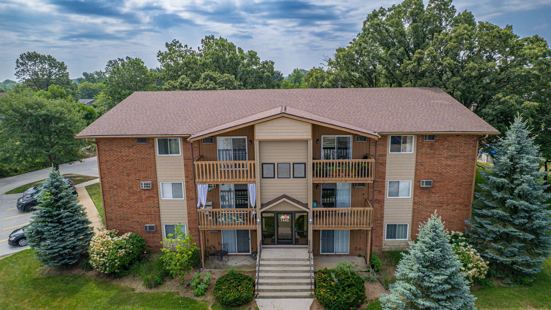 Apartment building with brick exterior, balconies, brown roof, and surrounding trees.