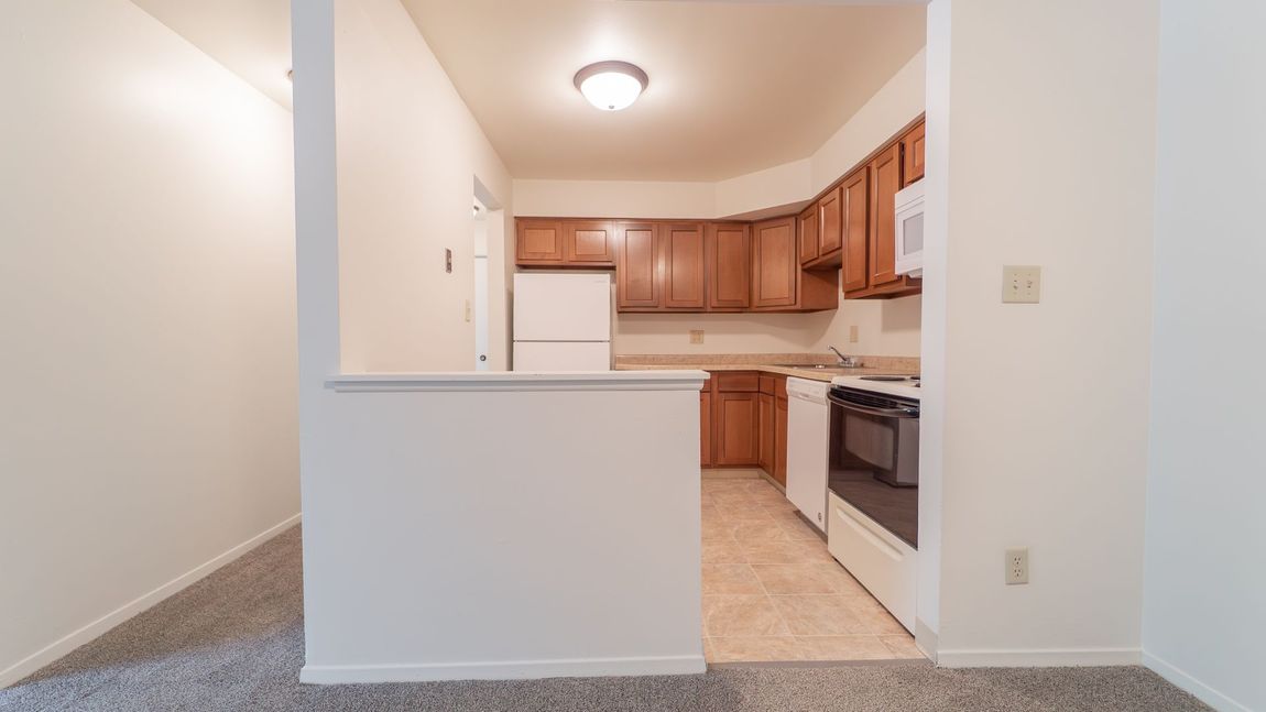 Kitchen with wooden cabinets, white appliances, and a half wall. Beige flooring and white walls.