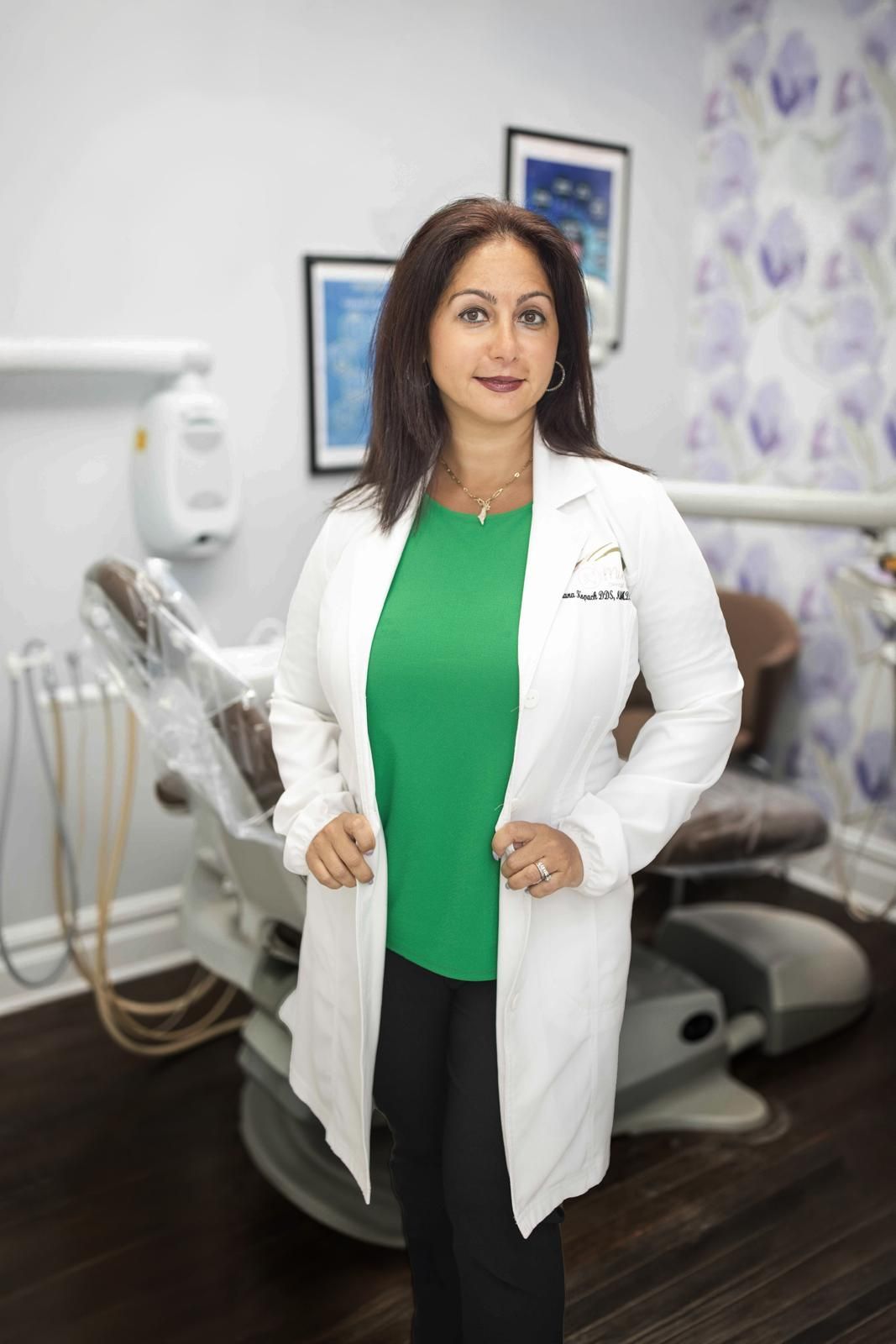 Woman in white lab coat and green shirt, standing in a dental office, smiling.