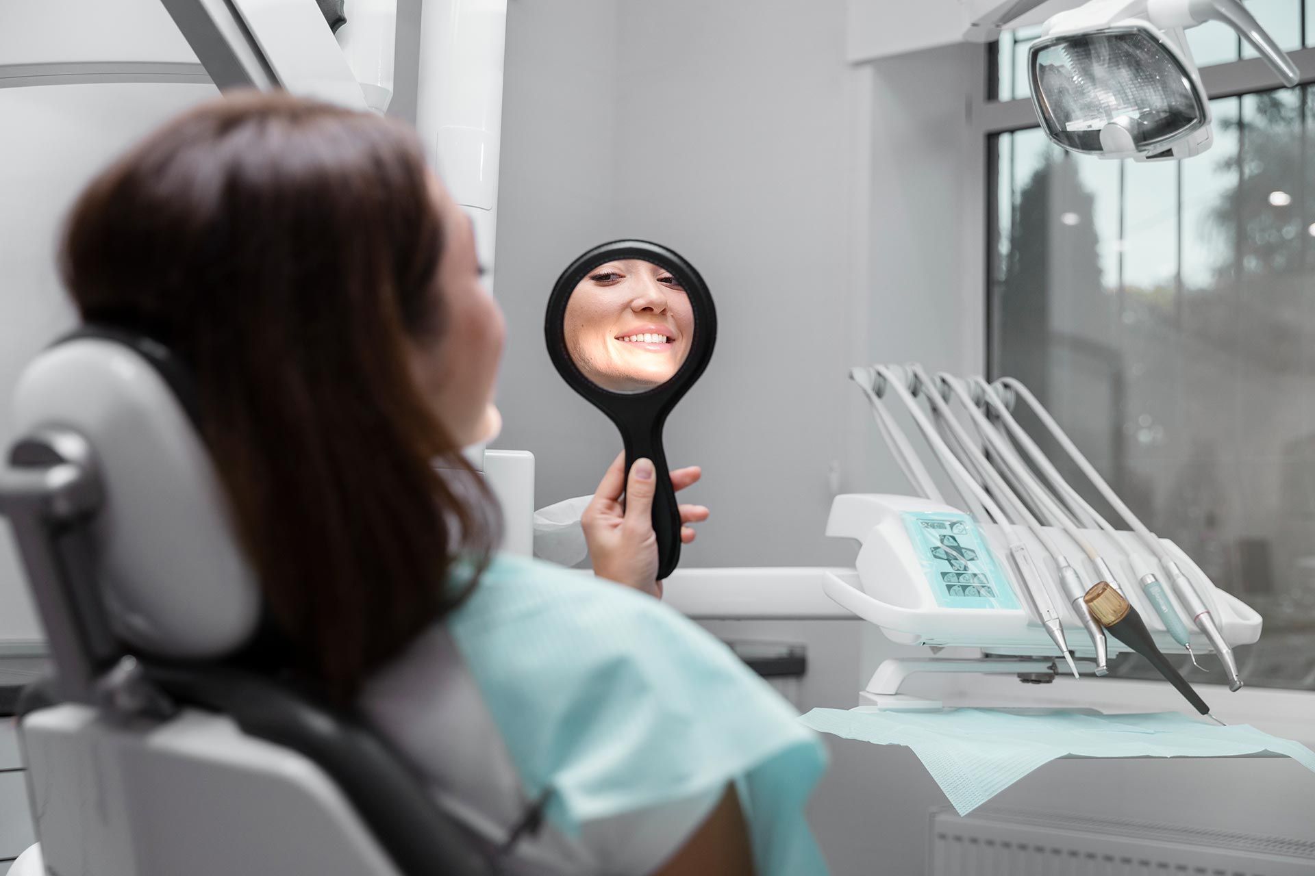 A woman is sitting in a dental chair looking at her smile in a mirror.