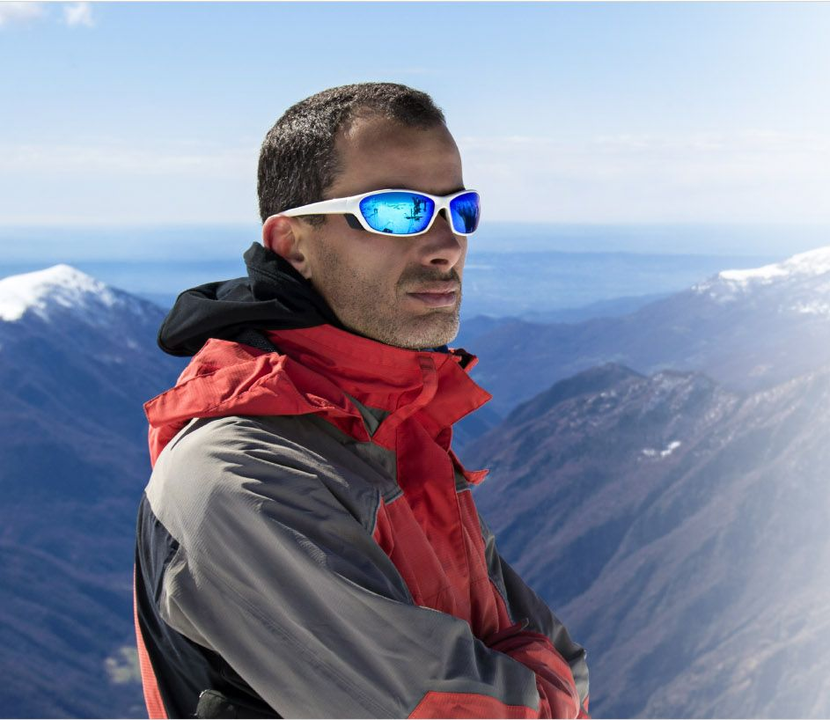 Escursionista con giacca rossa e occhiali da sole blu in piedi su una cresta montuosa innevata con cime lontane alle sue spalle