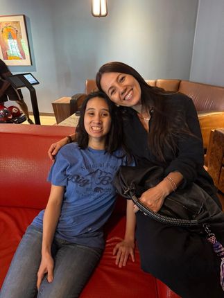 Two people smiling, seated on a red couch. One wears a blue shirt and jeans, the other has a black outfit, and a purse.