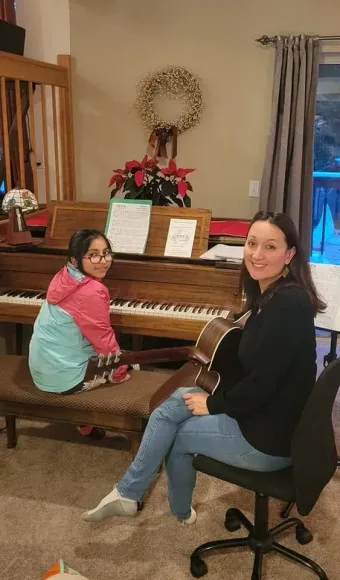 Two people at a piano, one showing sheet music to the other in a bright room.