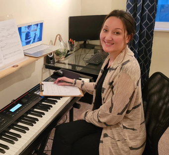 Two people sit at a piano, one playing while the other reads sheet music in a cozy living room.