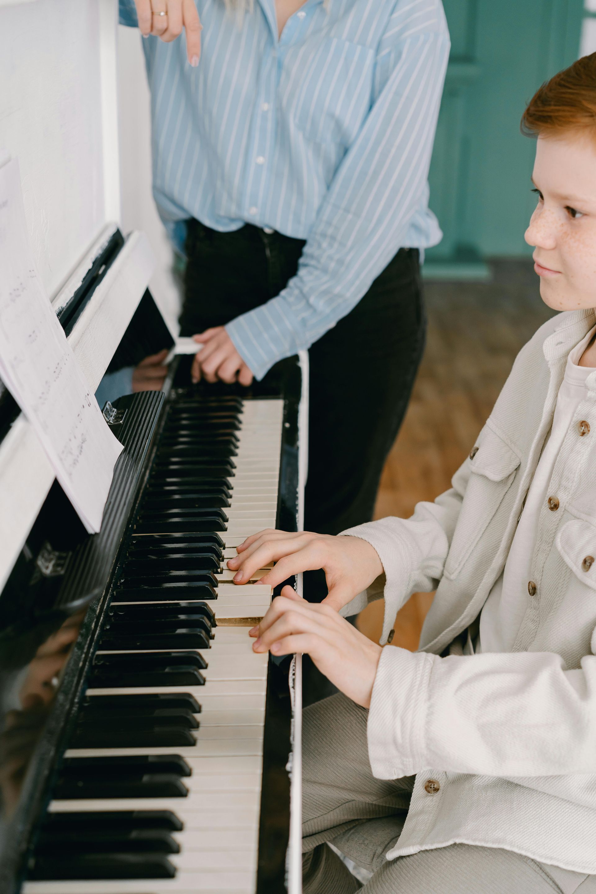 Two people play a piano together in a bright room, one seated and one standing beside the keys.