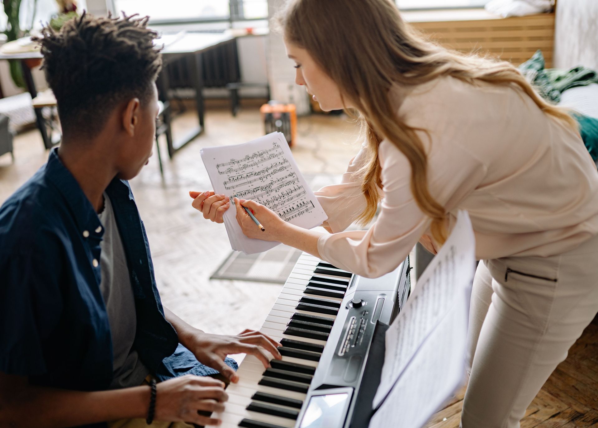 Teacher pointing at sheet music while a student plays keyboard in a bright room