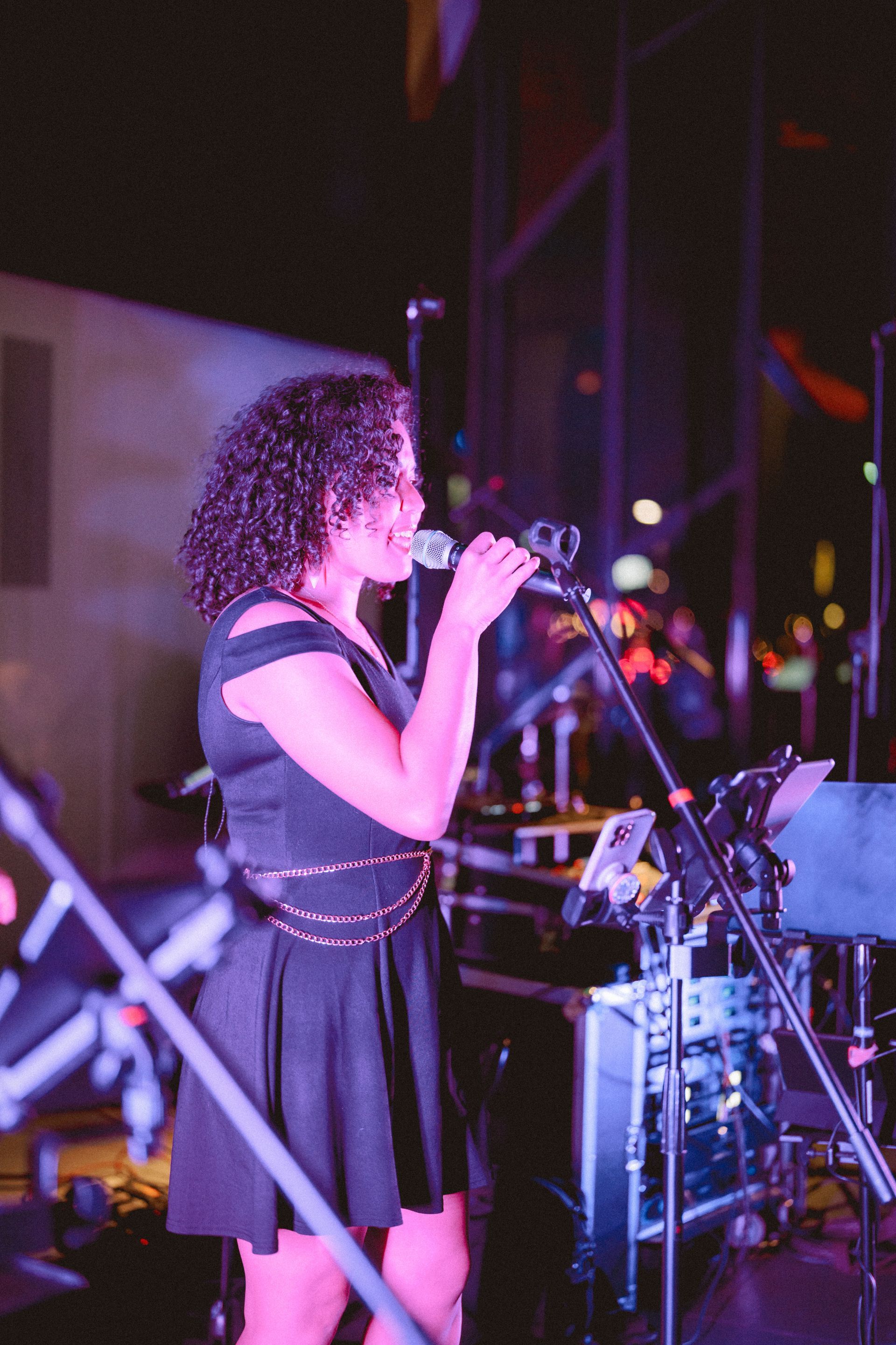 Drummer playing on stage with purple stage lights. Another musician in the background. Outdoor setting.