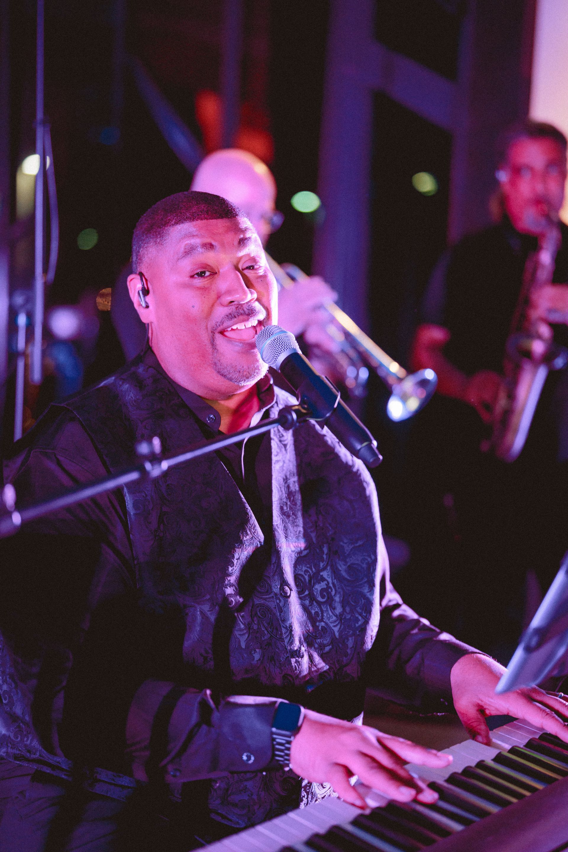 Band performing on an outdoor stage at dusk. Keyboardist, saxophone player, and singers. Green stage lighting.