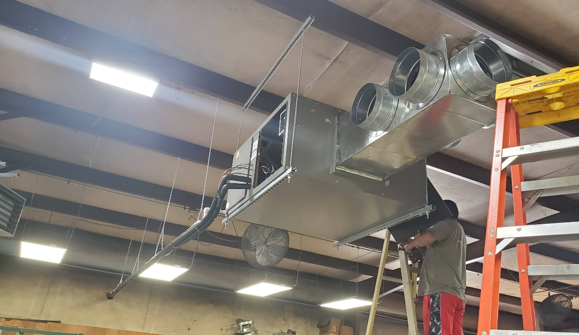 HVAC ductwork installation in a building. A person on a ladder works on hanging metal ductwork.
