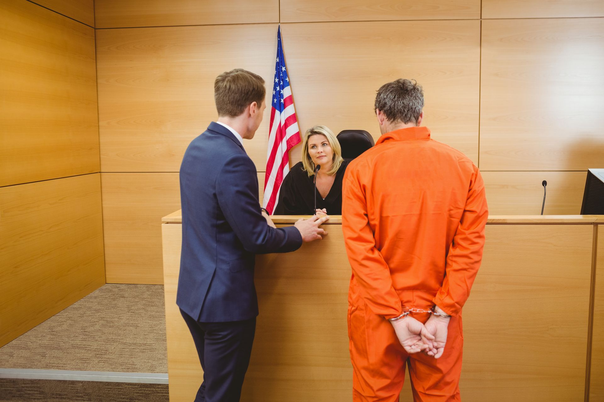 Attorney and handcuffed defendant standing before a judge in a courtroom with a U.S. flag.