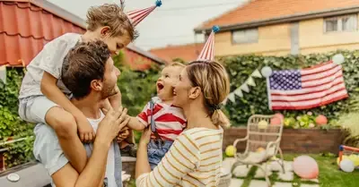 Family celebrating outdoors with American flags; man carries a child while two others smile in the yard