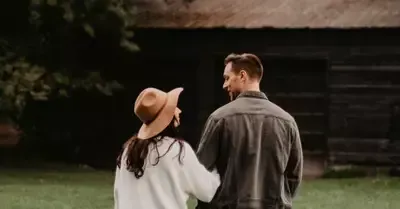 Two people stand outdoors, one in a tan hat and white sweater, facing a man in a gray jacket near a dark building.