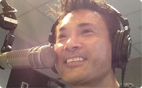 Person speaking into a studio microphone while wearing headphones and smiling.