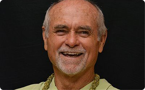 Smiling man with gray hair and beard on a dark background, wearing a light shirt and gold necklace