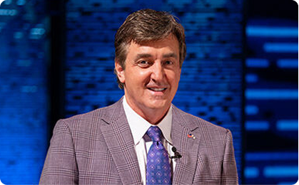 Man in a plaid blazer and blue tie smiling on a blue-lit stage