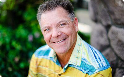 Smiling man in a yellow and blue shirt outdoors by greenery and a rock wall