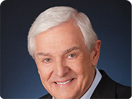 Smiling man with white hair in a dark suit against a blue studio background