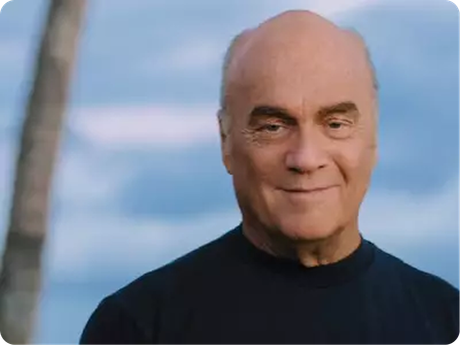 Portrait of a bald man in a dark shirt, smiling slightly against a blurred blue sky backdrop
