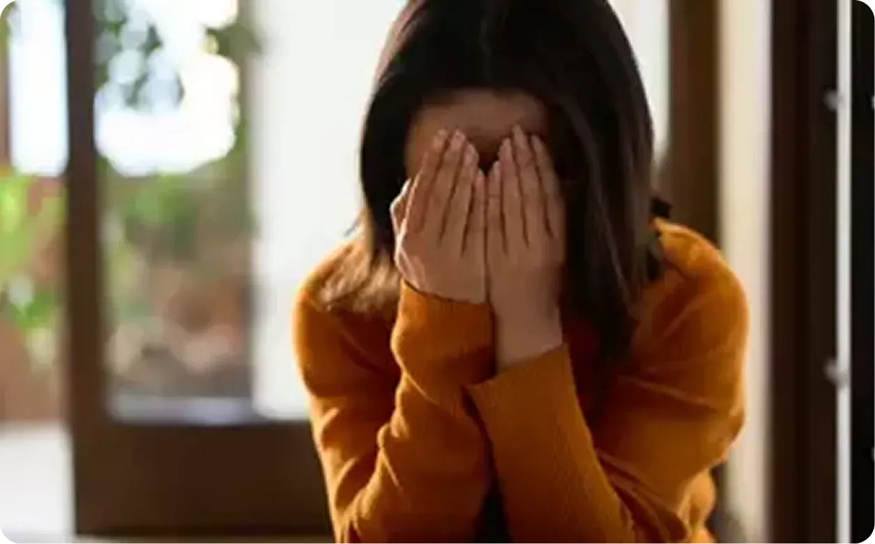 Person in an orange sweater sits indoors with face covered by both hands.