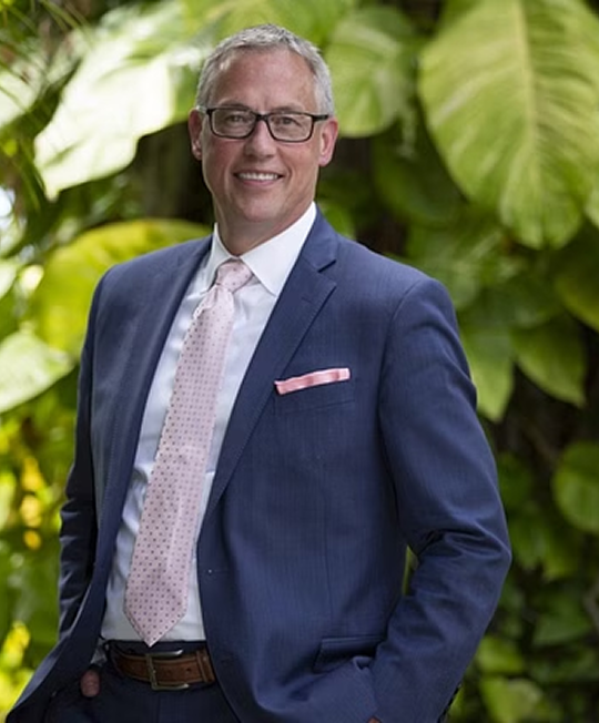 Man in blue suit, smiling, standing in front of greenery; wearing glasses and a pink tie with pocket square.