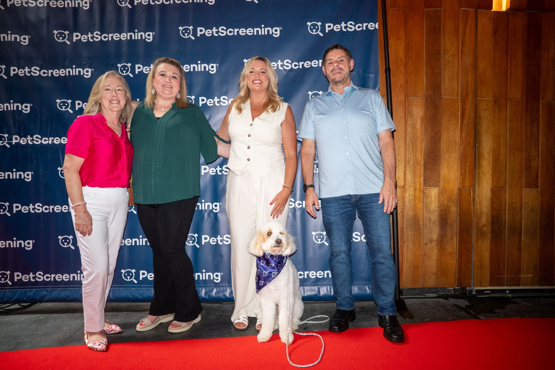Four people posing on a red carpet with a white dog in front of a PetScreening backdrop.