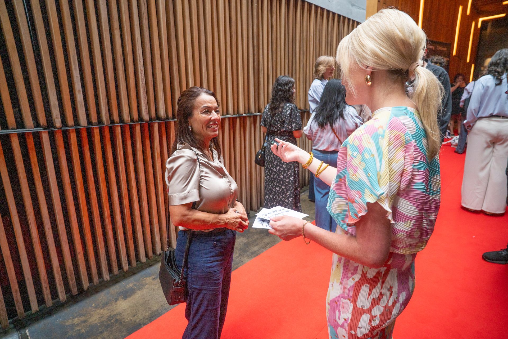 Two people talking on a red-carpet walkway, one holding a microphone, with vertical wood slats in the background.