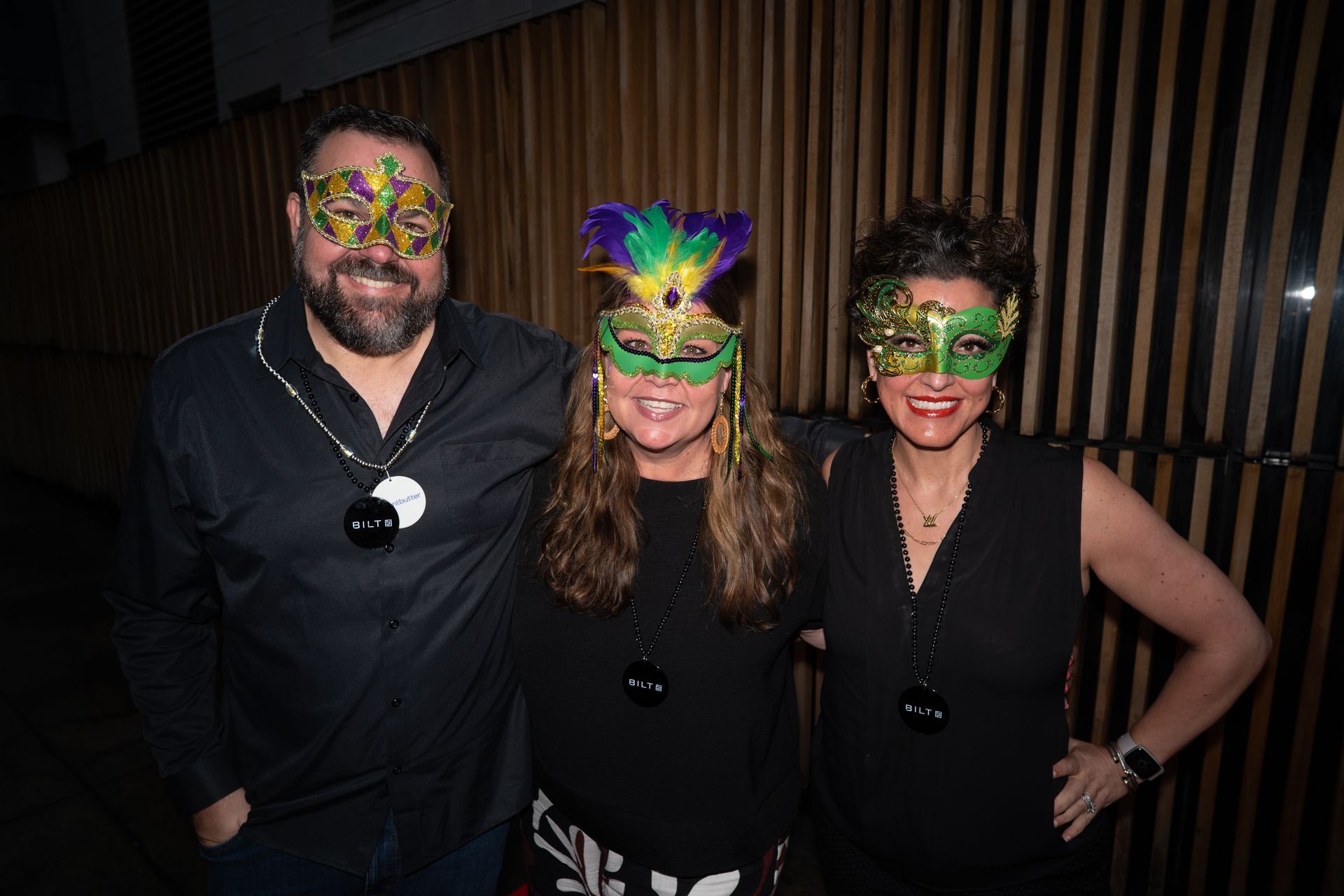 Three people wearing colorful masks pose together at a party in front of a dark wood wall.