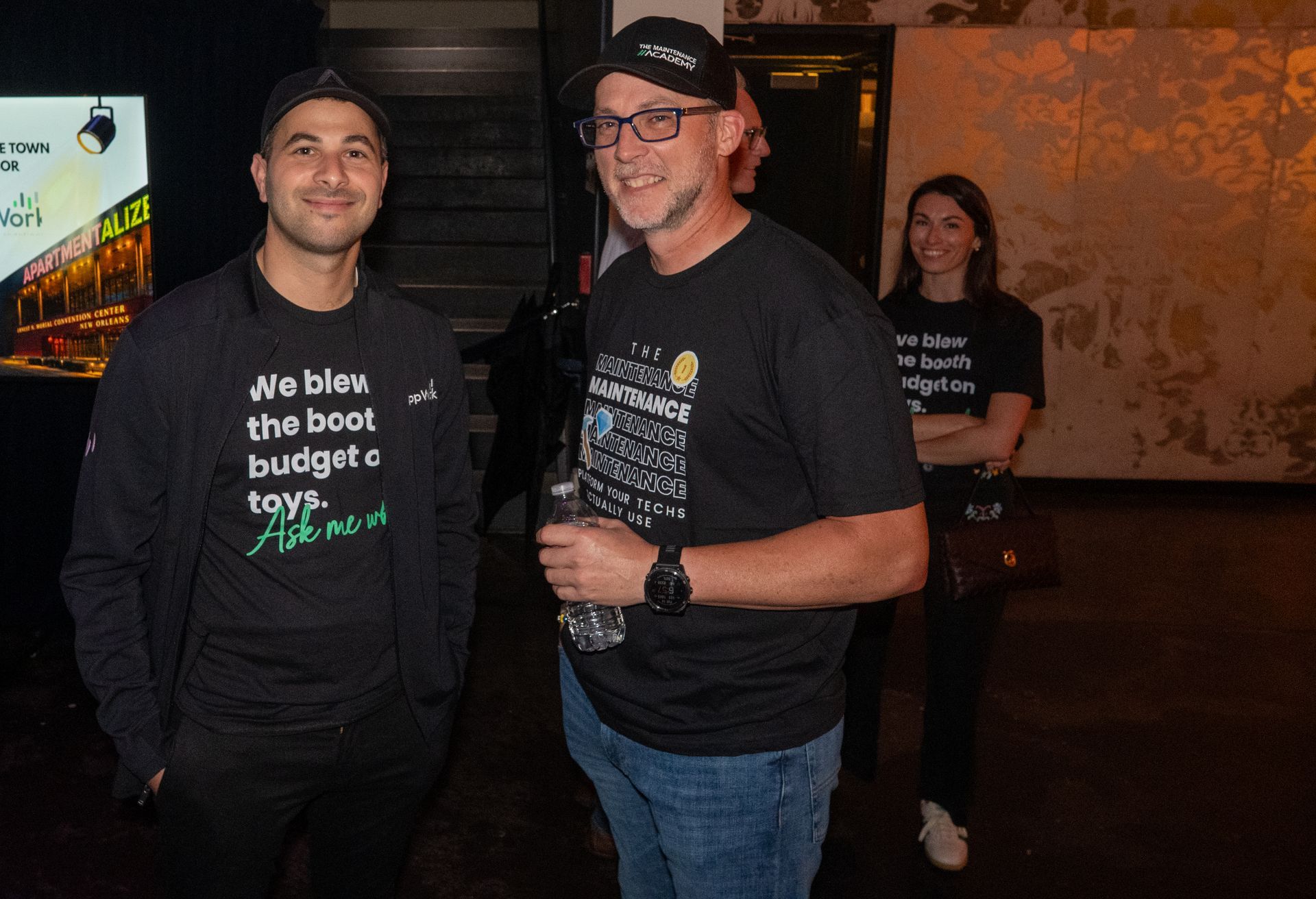 Two men in black T-shirts stand smiling indoors at a dim event venue.