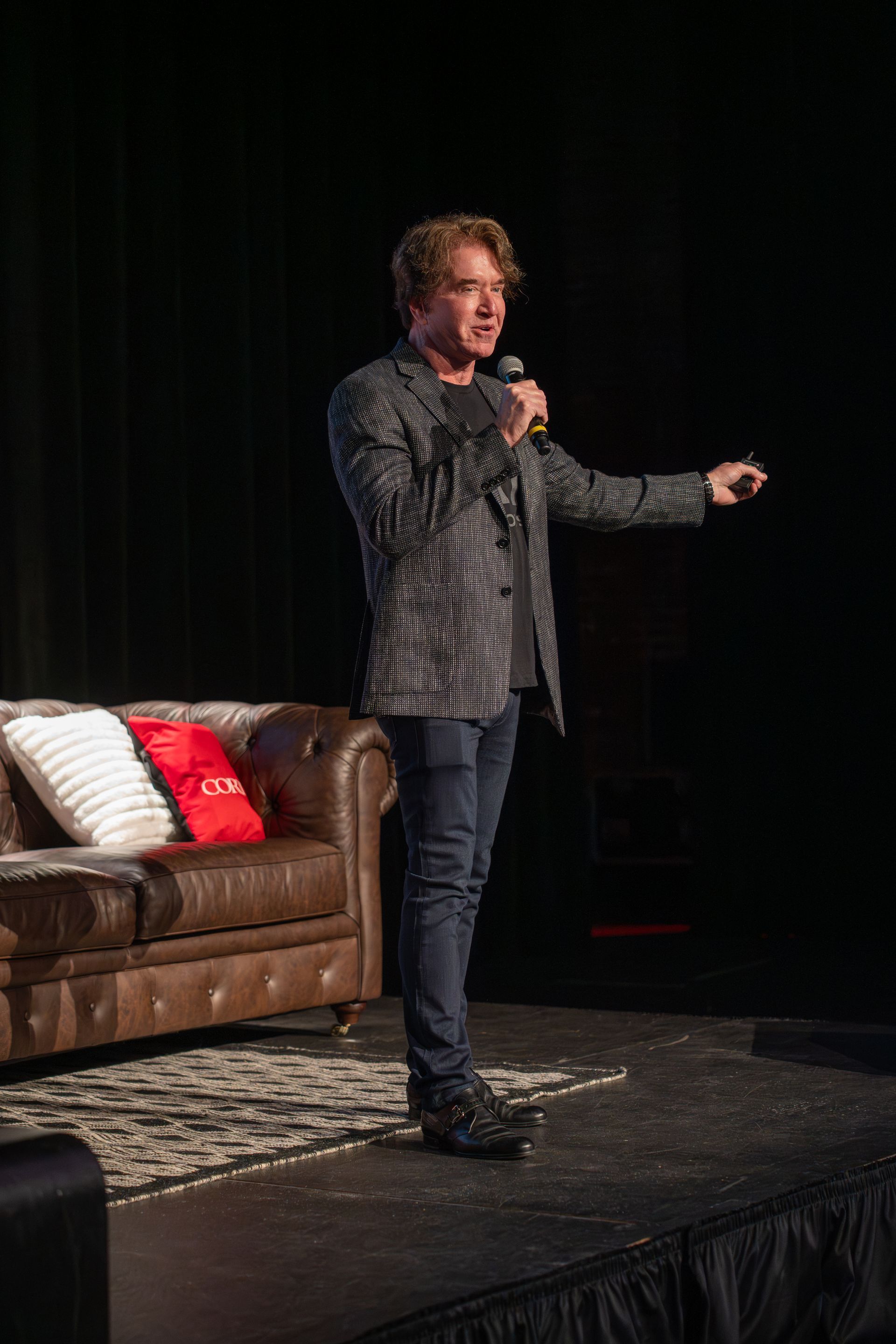 Kerry Kirby onstage holding a microphone beside a couch, speaking under a spotlight.