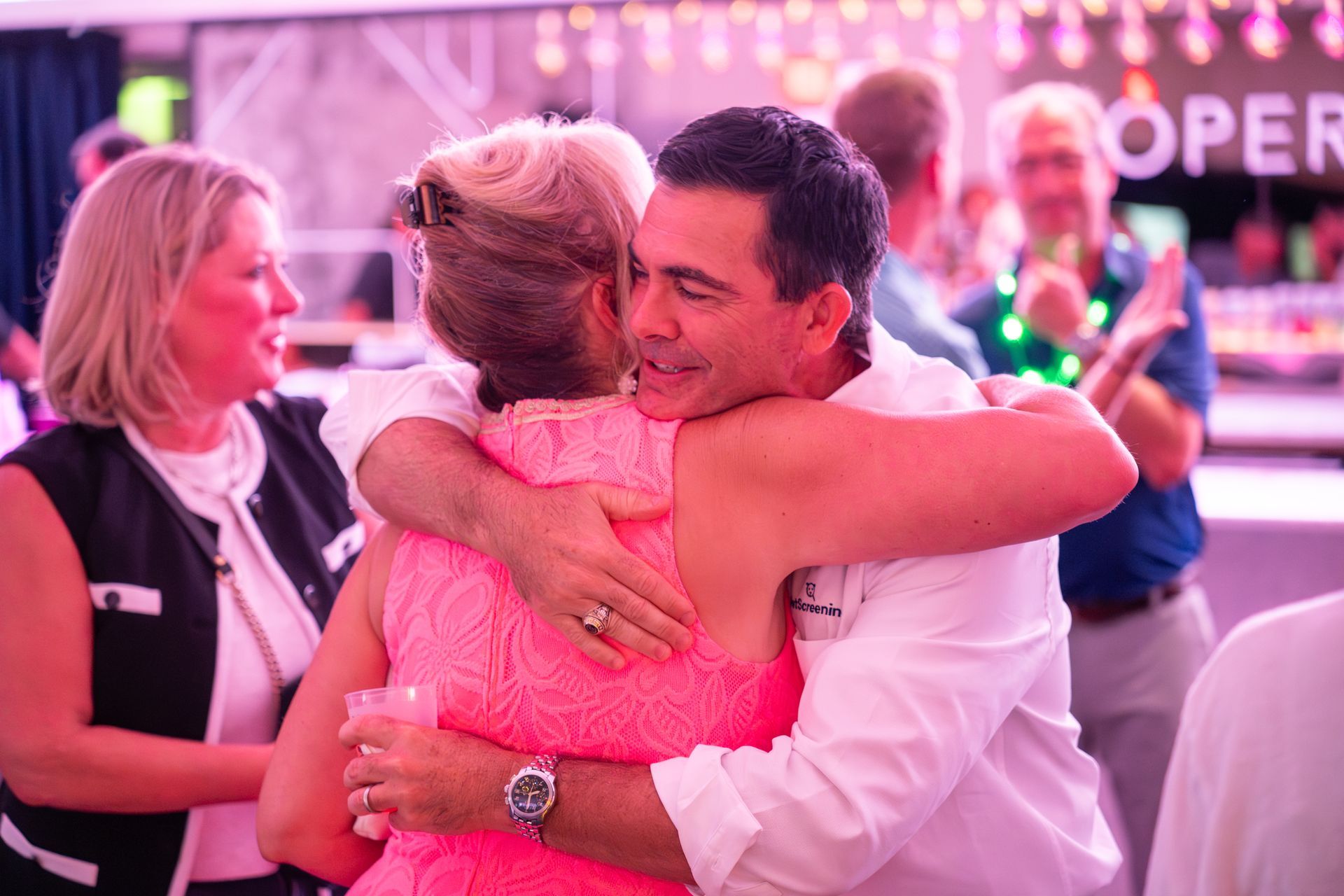Two people in pink embrace at a lively indoor event with other guests nearby.