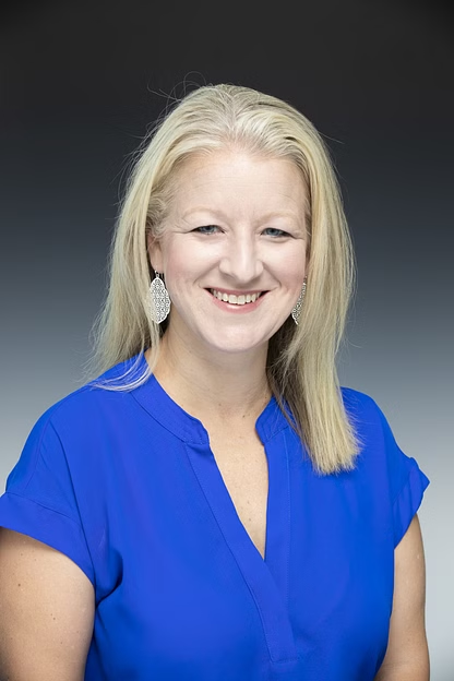 Woman with blonde hair, smiling, in a light blue collared shirt.