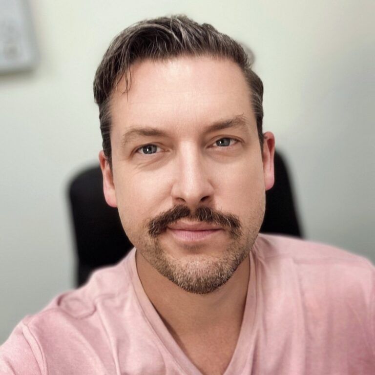 Man with mustache, light skin, looking at the camera, wearing a pink shirt.