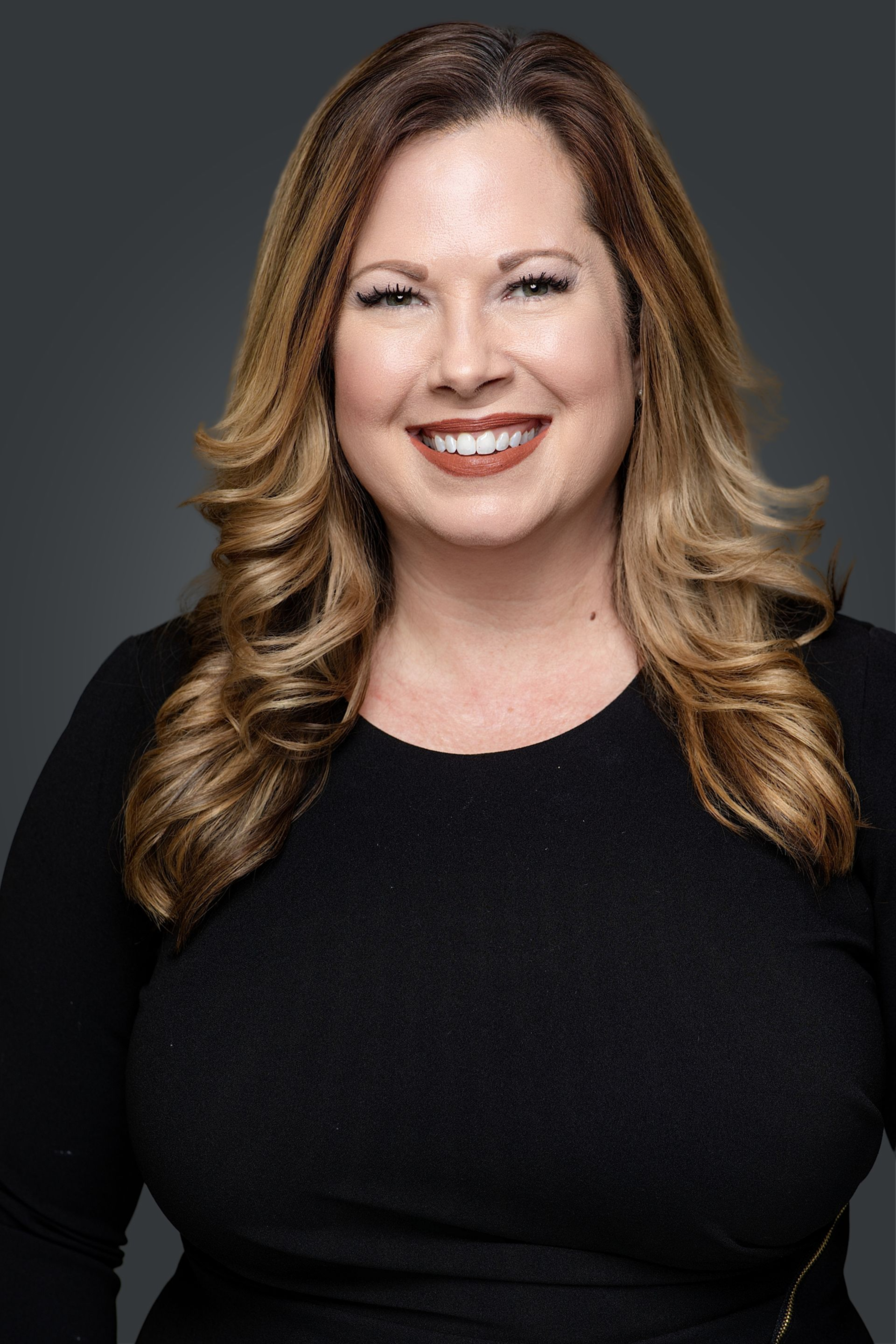 Woman with shoulder-length brown hair, smiling with red lipstick, wearing a black top against a gray backdrop.