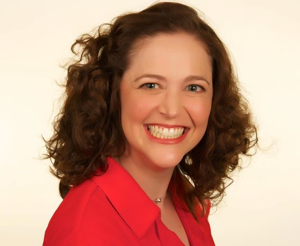 Woman with curly brown hair smiles brightly, wearing a red top, against a pale yellow background.
